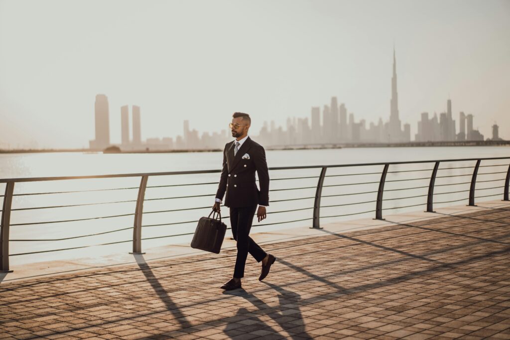 Professional man in formal suit walking by Dubai waterfront with city skyline and Burj Khalifa in view.